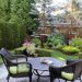 Black wicker patio chairs with green cushions surround a round table overlooking a lush backyard garden with flowers.