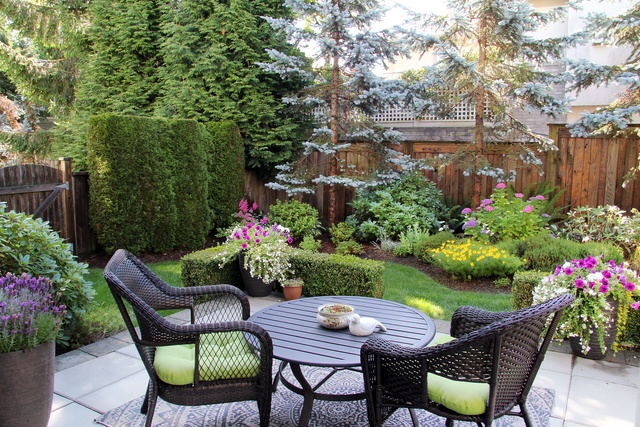 Black wicker patio chairs with green cushions surround a round table overlooking a lush backyard garden with flowers.