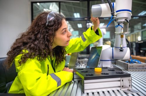 A worker in a high-visibility jacket inspecting a robotic arm and equipment at an industrial workstation.