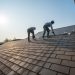 Two roofers wearing safety harnesses work on a large, sloped shingle roof under a bright blue sky on a sunny day.