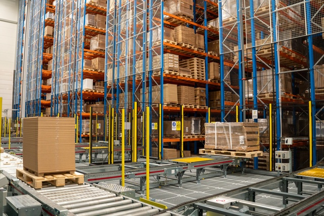 Pallets stacked with cardboard boxes sit on roller conveyors inside a warehouse with tall blue and orange storage racks.