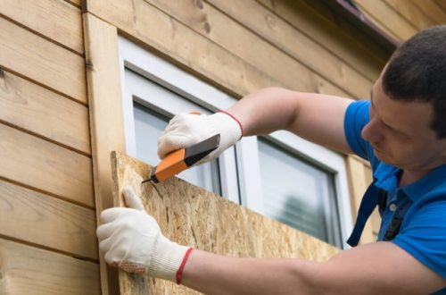 A person wearing work gloves and a blue shirt is using a hammer to attach a piece of wood in front of a window.