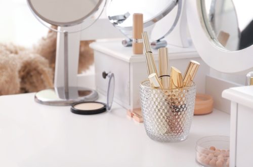 Gold-toned makeup brushes and cosmetic tools stand in a textured glass holder on a bright white vanity with mirrors.