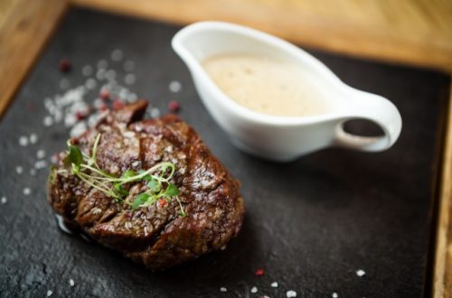 A cooked filet mignon sits on a black surface next to a white gravy boat filled with a cream-colored liquid.